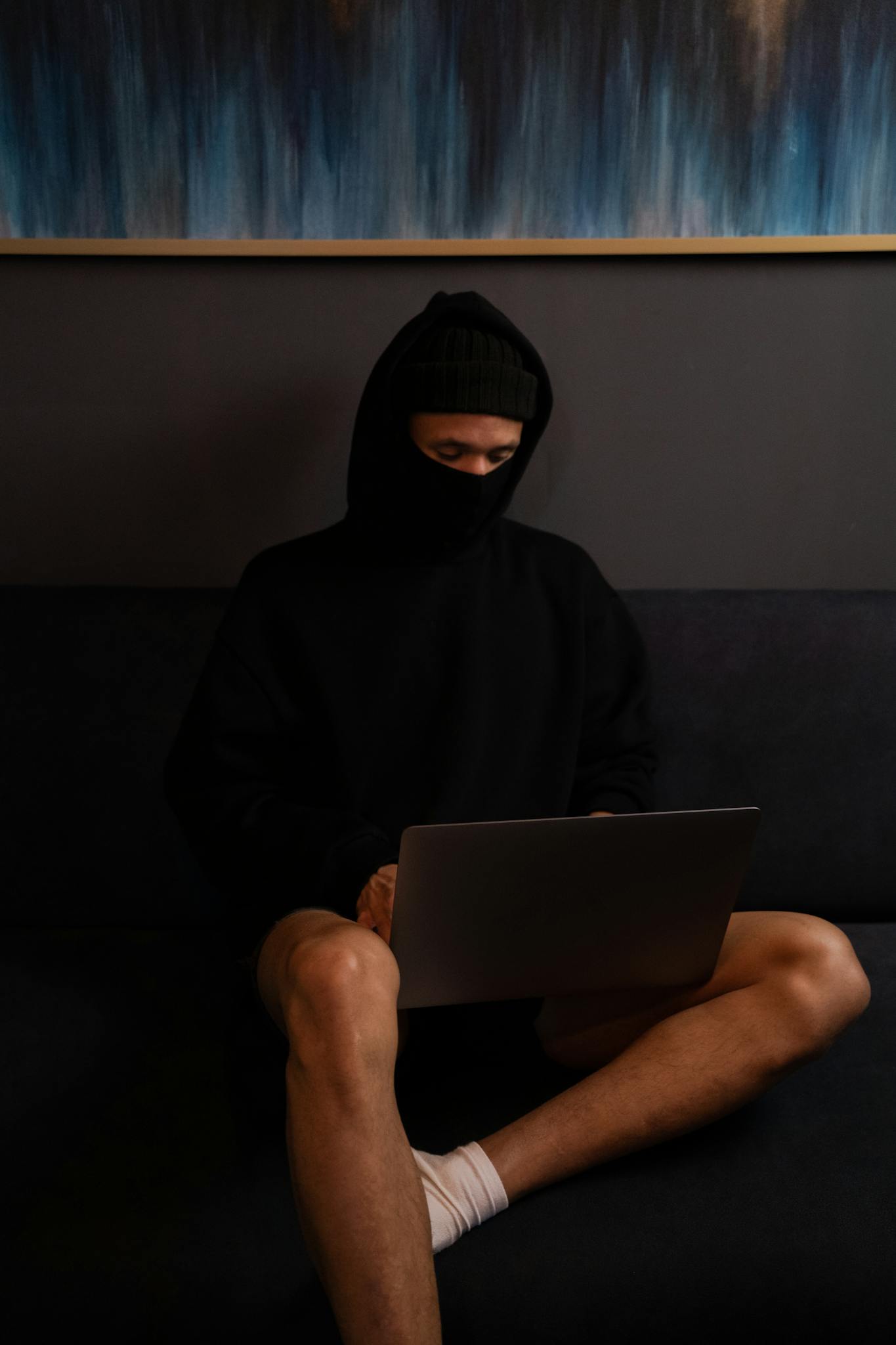 An individual sitting indoors with a laptop, wearing dark clothing and a face mask.
